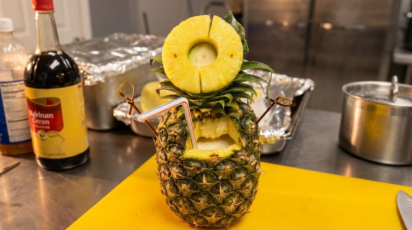 A carved pineapple drink with a straw and pineapple slice garnish on a yellow cutting board.