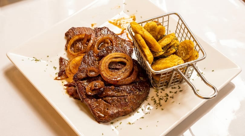 Steak with caramelized onions and a side of fried plantains in a metal basket.