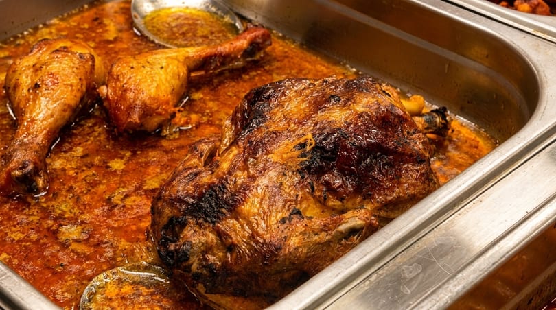 Roasted whole chicken and drumsticks in a metal pan with rich orange sauce.